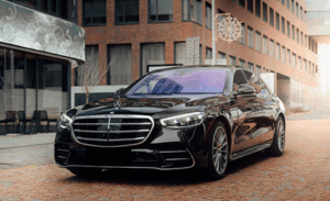 A black luxury sedan, likely a Mercedes-Benz, is parked on a cobblestone street in an urban setting, with modern buildings in the background.
