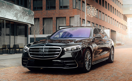 A black luxury sedan, likely a Mercedes-Benz, is parked on a cobblestone street in an urban setting, with modern buildings in the background.
