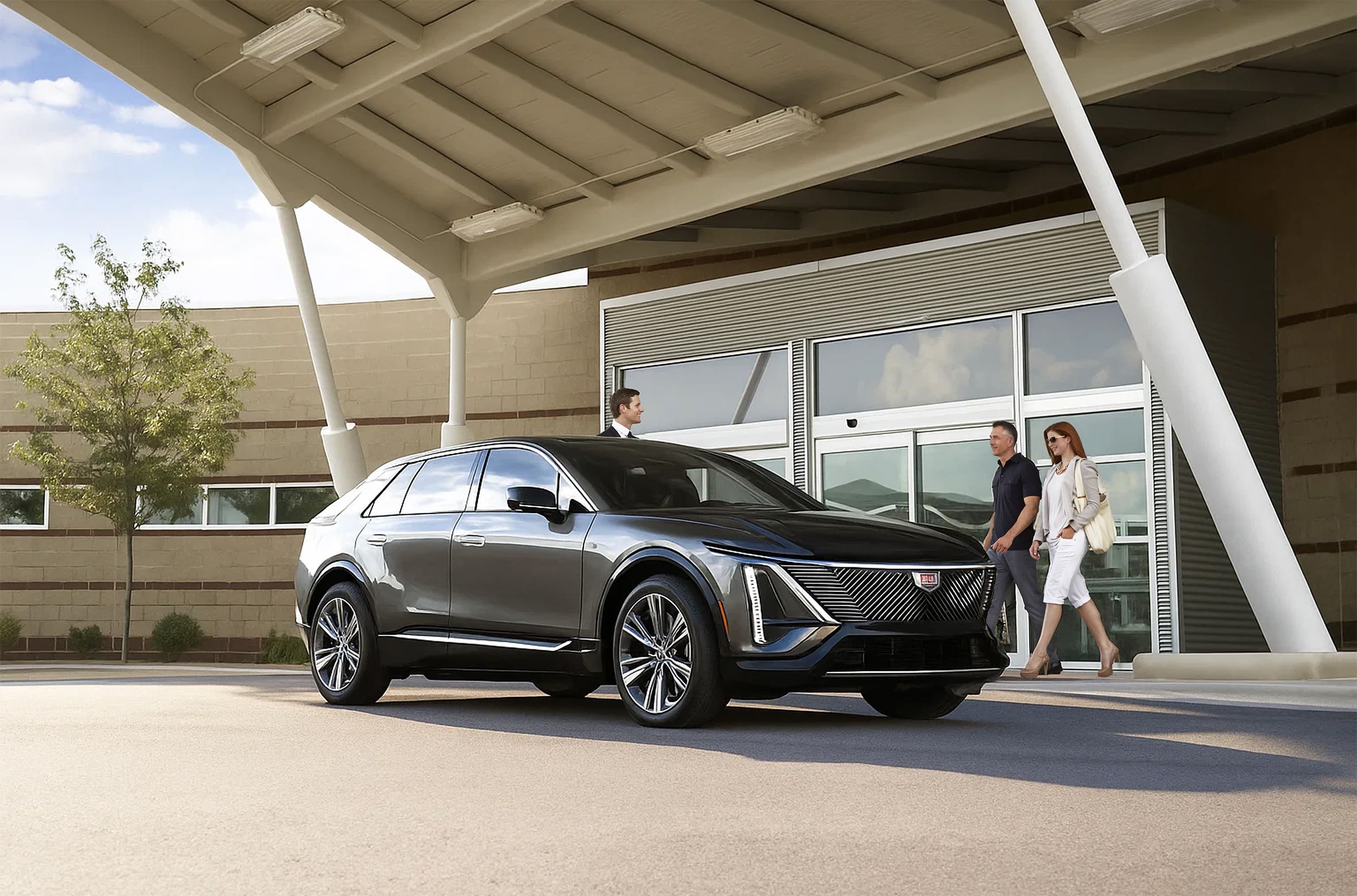 Carey chauffeur behind a black Cadillac Lyriq at the airport, preparing to open the door for two passengers coming from baggage claim.